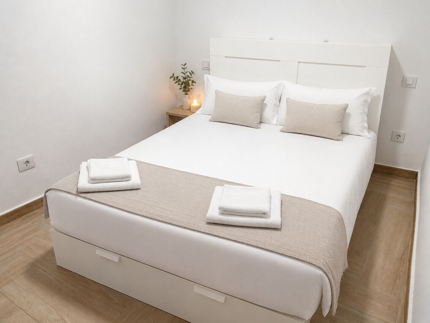 A neatly made bed with white linens and a beige throw blanket, a bedside table with a candle, and a small plant.