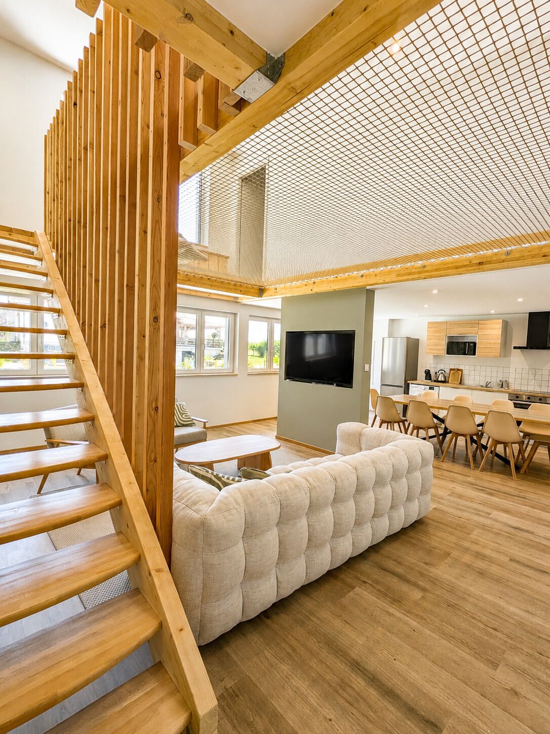 Een modern interieur met een houten trap, een zithoek met bank en salontafel, een keuken met eethoek, en een grote televisie aan de muur.