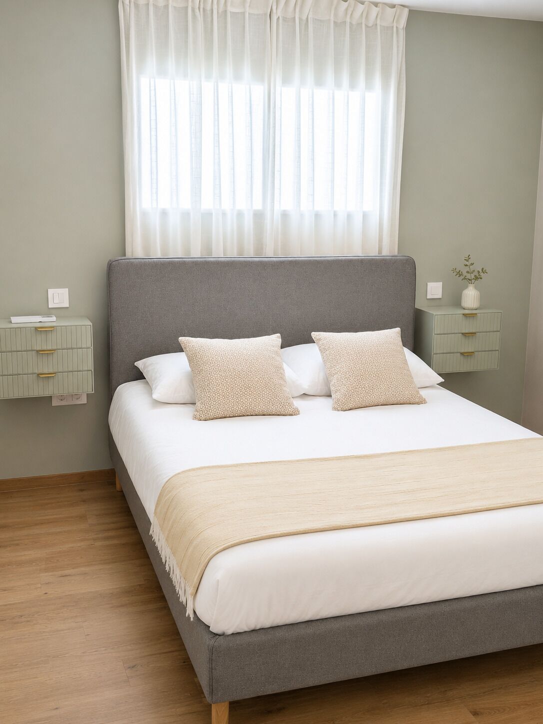 A neatly made bed with a grey headboard, white bedding, and two beige pillows. There are two bedside tables with decorative vases, and a window with sheer curtains.