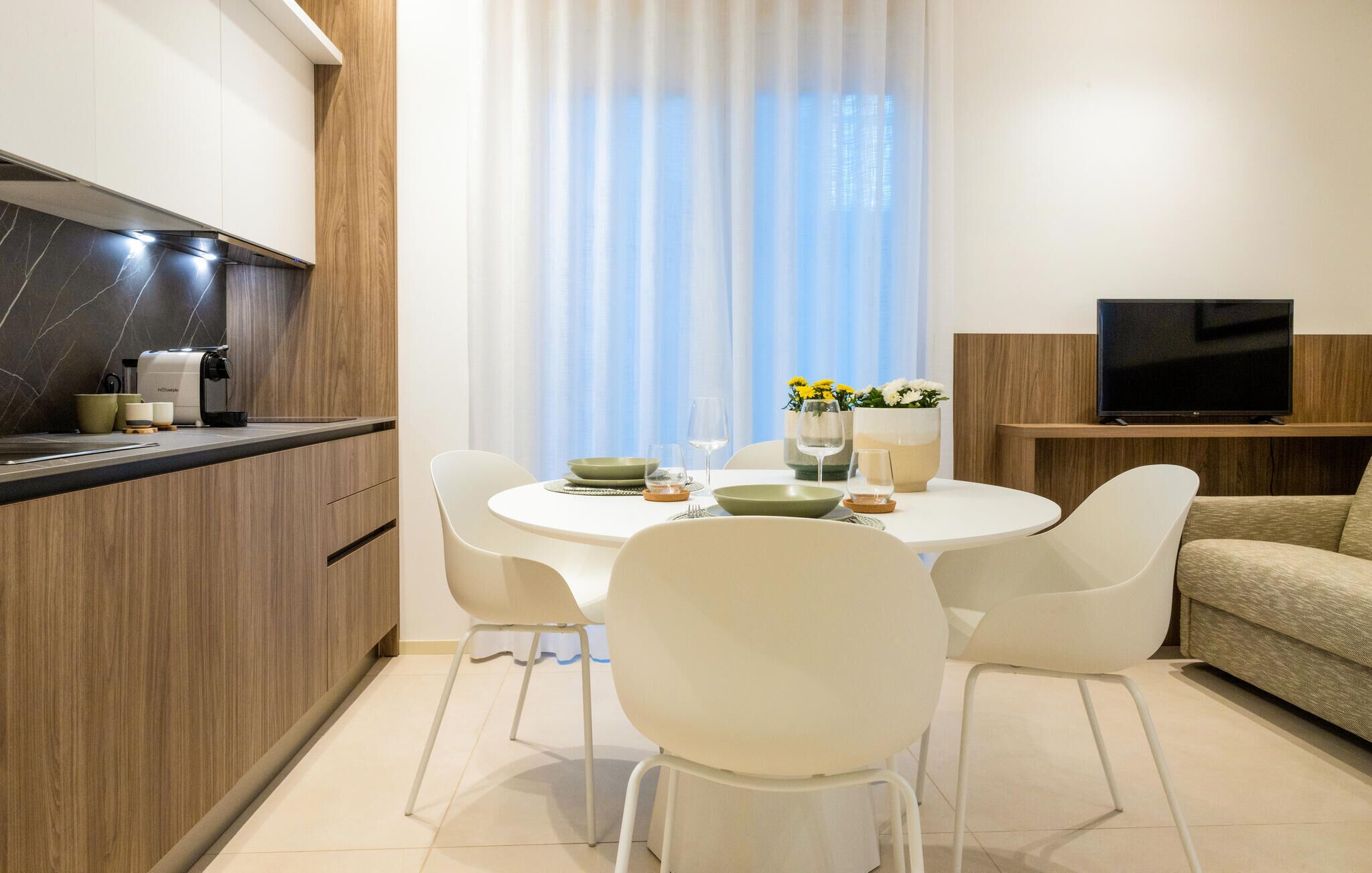 A modern kitchen with a built-in stove, white chairs, a dining table set for two, and a TV on a wooden stand.