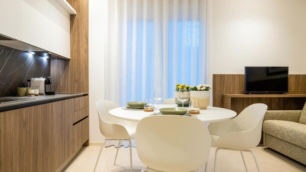 A modern kitchen with a built-in stove, white chairs, a dining table set for two, and a TV on a wooden stand. - Cozy apartment in Marina di Ragusa with house a panoramic view (Marina di Ragusa)