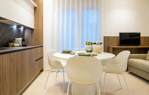 A modern kitchen with a built-in stove, white chairs, a dining table set for two, and a TV on a wooden stand. - Cozy apartment in Marina di Ragusa with house a panoramic view (Marina di Ragusa)