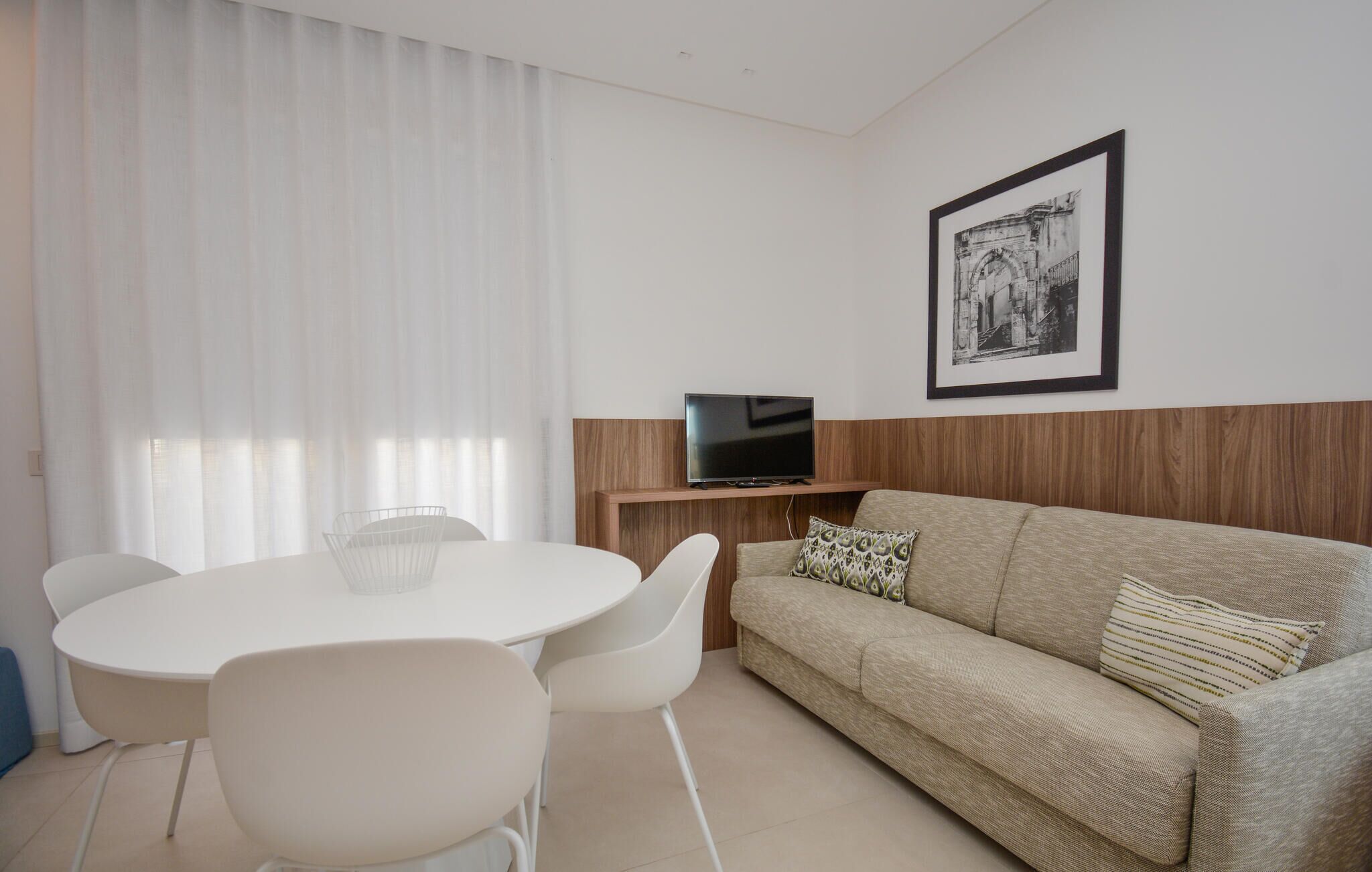 A modern living room with a round dining table, white chairs, a beige sofa, and a television on a wooden stand.
