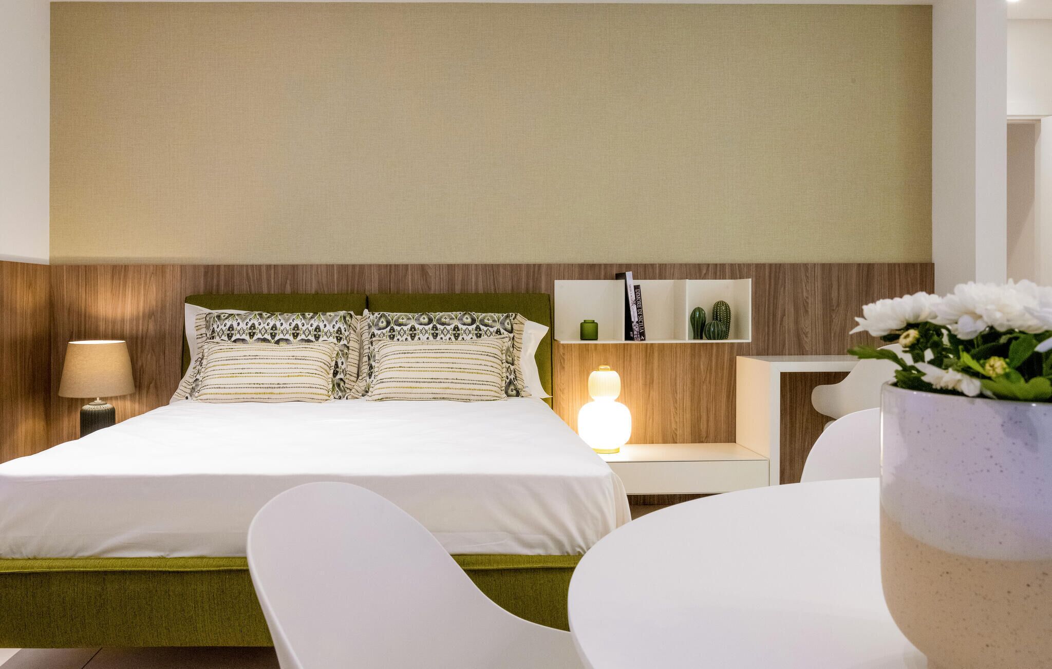 A hotel room with a bed, bedside table, lamp, and a small shelf with books and decorative items.