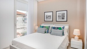 Ein Hotelzimmer mit einem Bett, zwei Nachttischen, einem Fenster mit Blick auf Gebäude und zwei gerahmten Bildern an der Wand.