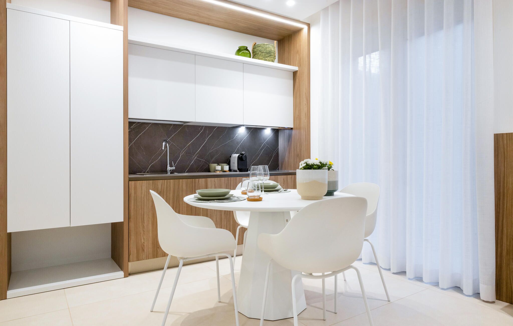 Una cocina moderna con armarios blancos, una salpicadera de mármol y un comedor con sillas blancas y una mesa redonda.