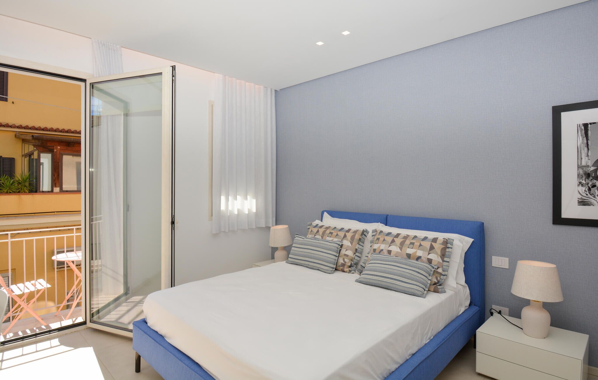 A bedroom with a bed, bedside lamps, a window with curtains, and a balcony with chairs.