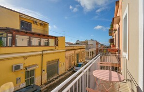 Eine schmale Straße mit Häusern an beiden Seiten, ein geparktes Auto und ein Balkon mit Tisch und Stühlen.