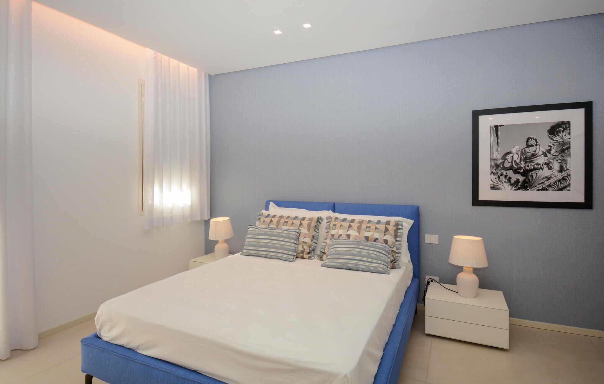 A bedroom with a bed, two bedside tables, a nightstand, a framed picture on the wall, and a window with curtains.