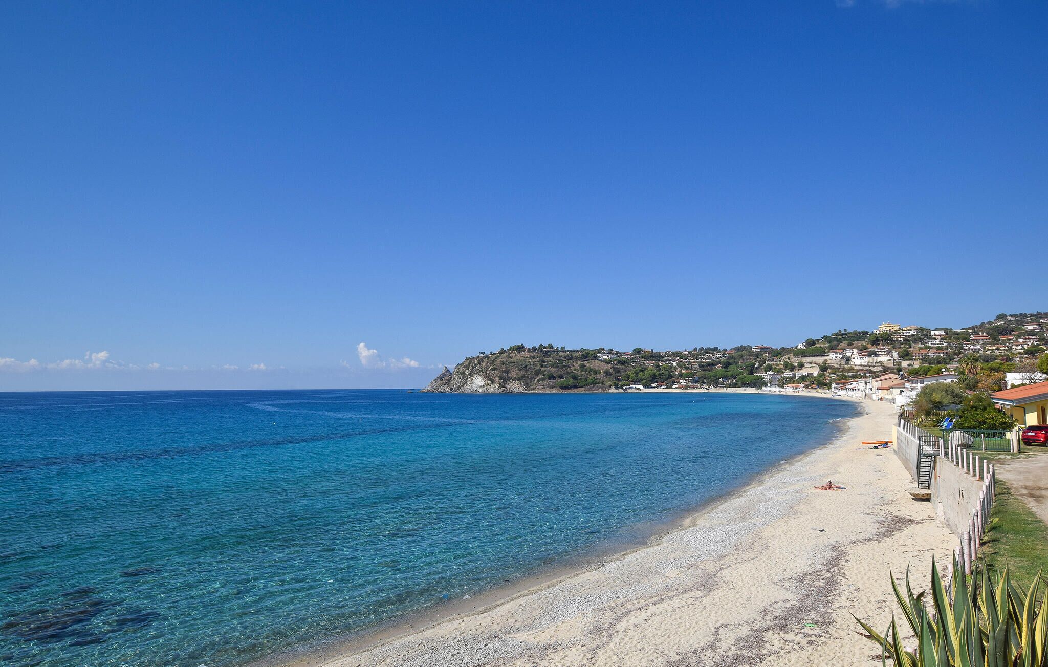 A coastal beach with clear blue waters, a sandy shore, and a hillside with buildings.