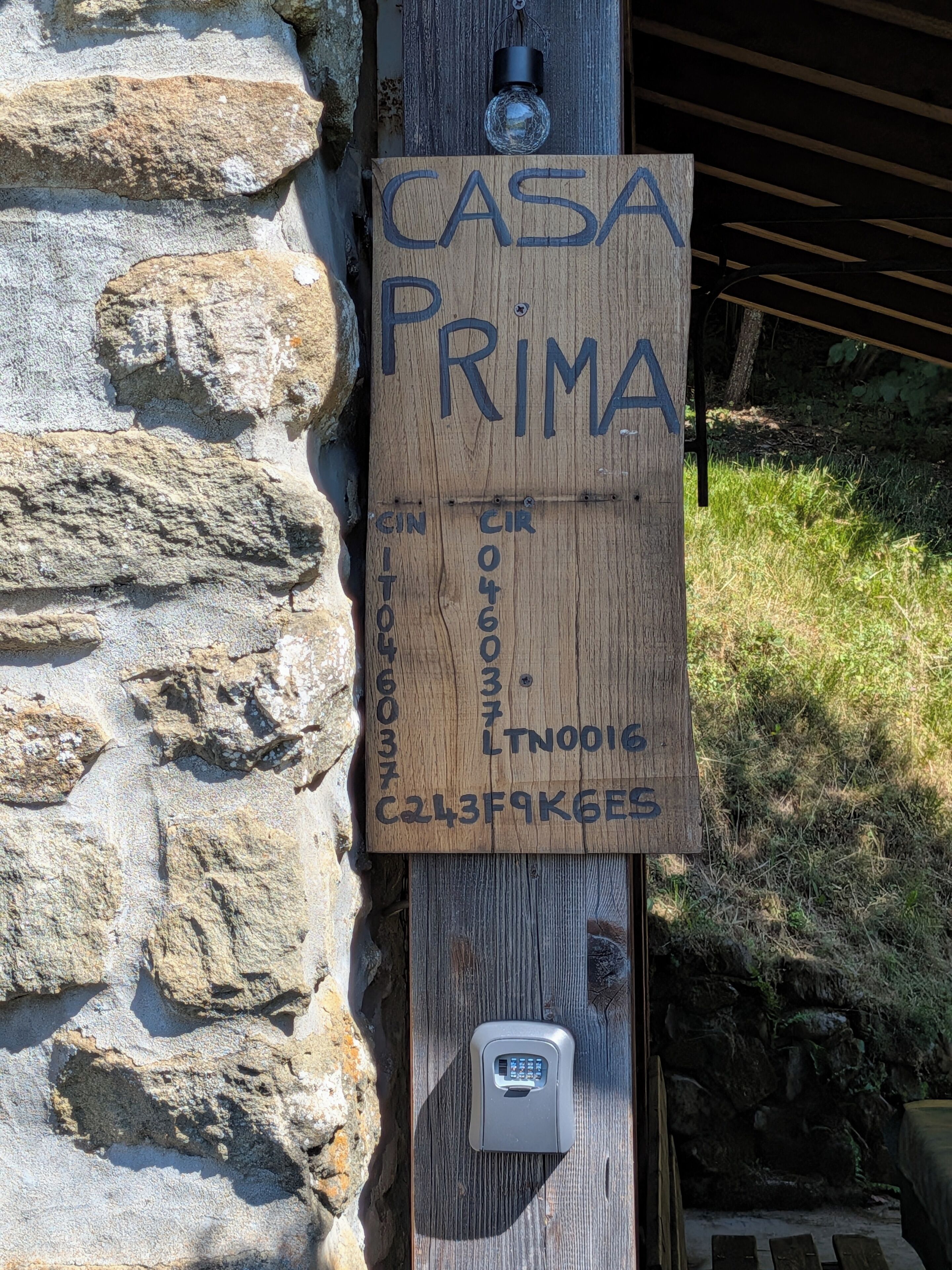 Ein hölzernes Schild mit der Aufschrift 'CASA PRIMA' und einer Telefonnummer, an einer Steinmauer befestigt.
