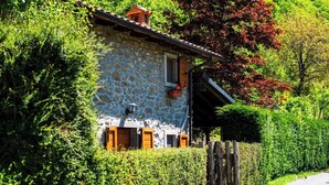 Une maison en pierre avec une porte en bois et des volets aux fenêtres, entourée d’une végétation luxuriante et d’une clôture en bois.