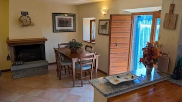 Zona pranzo con camino, sedie in legno, tavolo e bancone della cucina con un vaso di fiori.