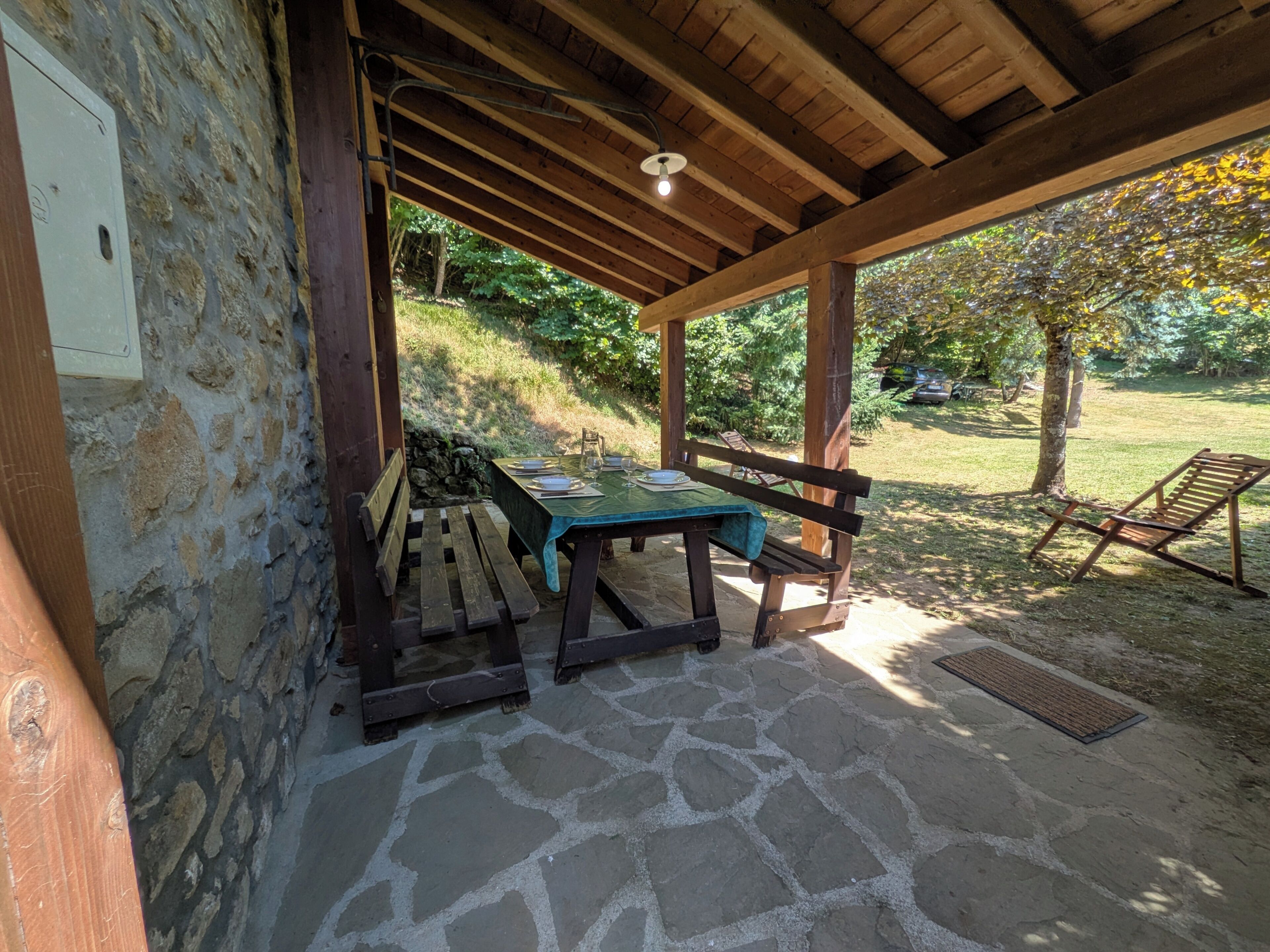 A covered outdoor seating area with a stone floor, wooden benches, and a table set for dining.