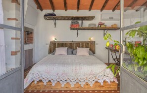 Ein Schlafzimmer mit einem hölzernen Kopfende, einem Bett mit einer Spitzenüberdecke, einem Regal mit verschiedenen Gegenständen und einer Topfpflanze.