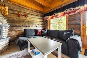 A log cabin interior with a stone fireplace, a sofa, a coffee table, and a window with a view of trees.