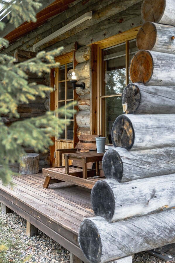 A rustic cabin with a wooden deck, a bench, a table, and a bucket.