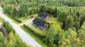 A log cabin with a black roof, surrounded by dense forest, with a paved road in the foreground.