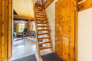 A wooden interior with a staircase leading to a second floor, a dining area with chairs and a table, and a view of the outdoors through a window.