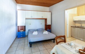 Ein kleines Schlafzimmer mit einem Bett, einem Tisch mit Tasse und Untertasse, einem Sessel und einer Küchenzeile mit Spüle und Schränken.
