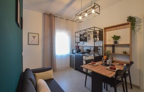A compact living space with a kitchenette, a dining area with a table and chairs, a sofa, and a wall-mounted shelf with plants. - Le Vele - Contro Fiocco (Rosolina)