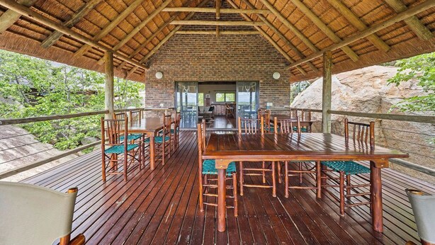 An outdoor wooden deck with a thatched roof, surrounded by greenery, and a view of a building in the background. - Boulders Bush Lodge: A Private Boulder-Top Retreat in Greater Kruger (Mica)