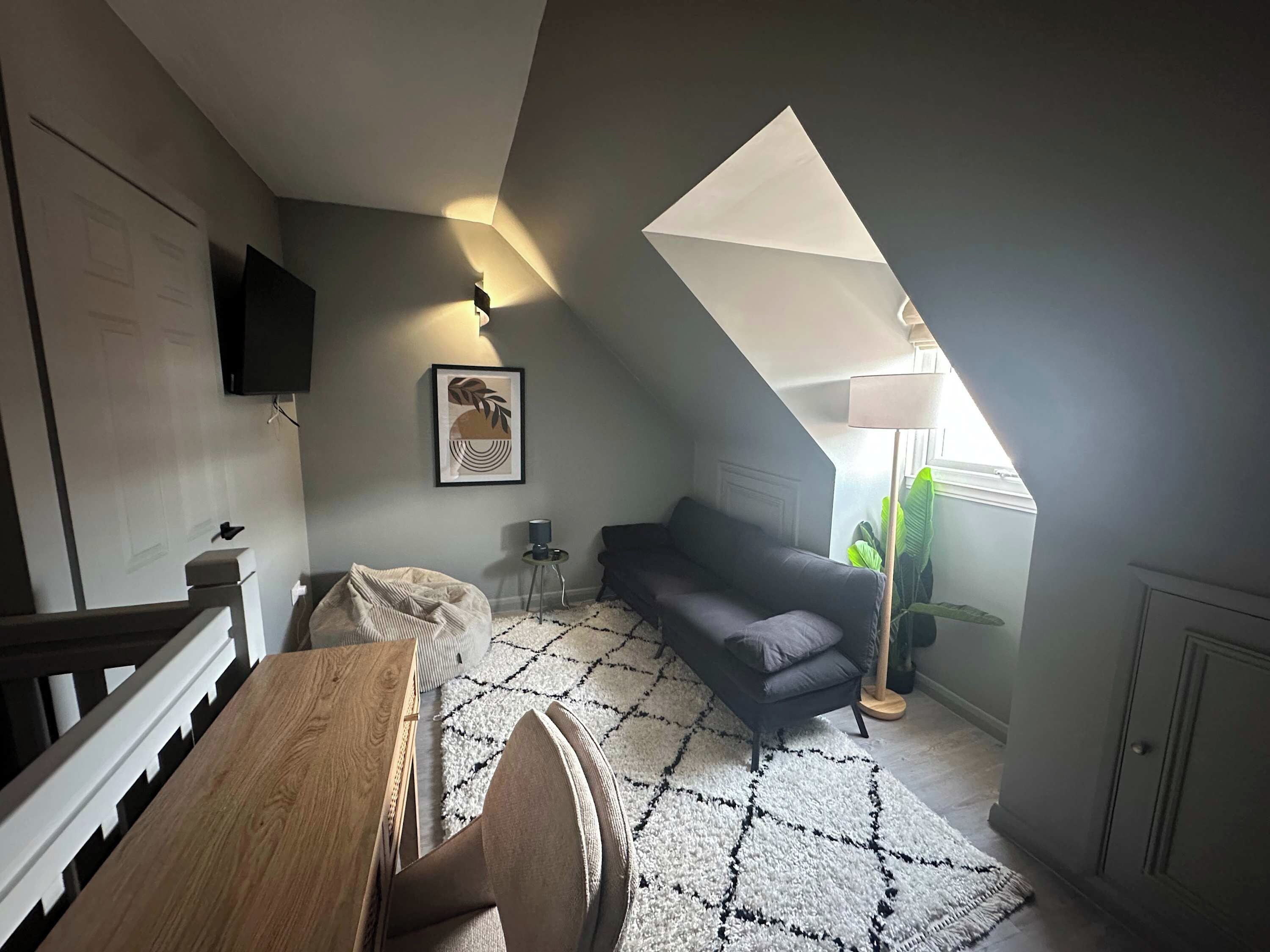 Un dormitorio estilo loft moderno con techo inclinado, una mesa de comedor de madera, un sofá gris oscuro, una alfombra estampada y un gran ventanal con luz natural.