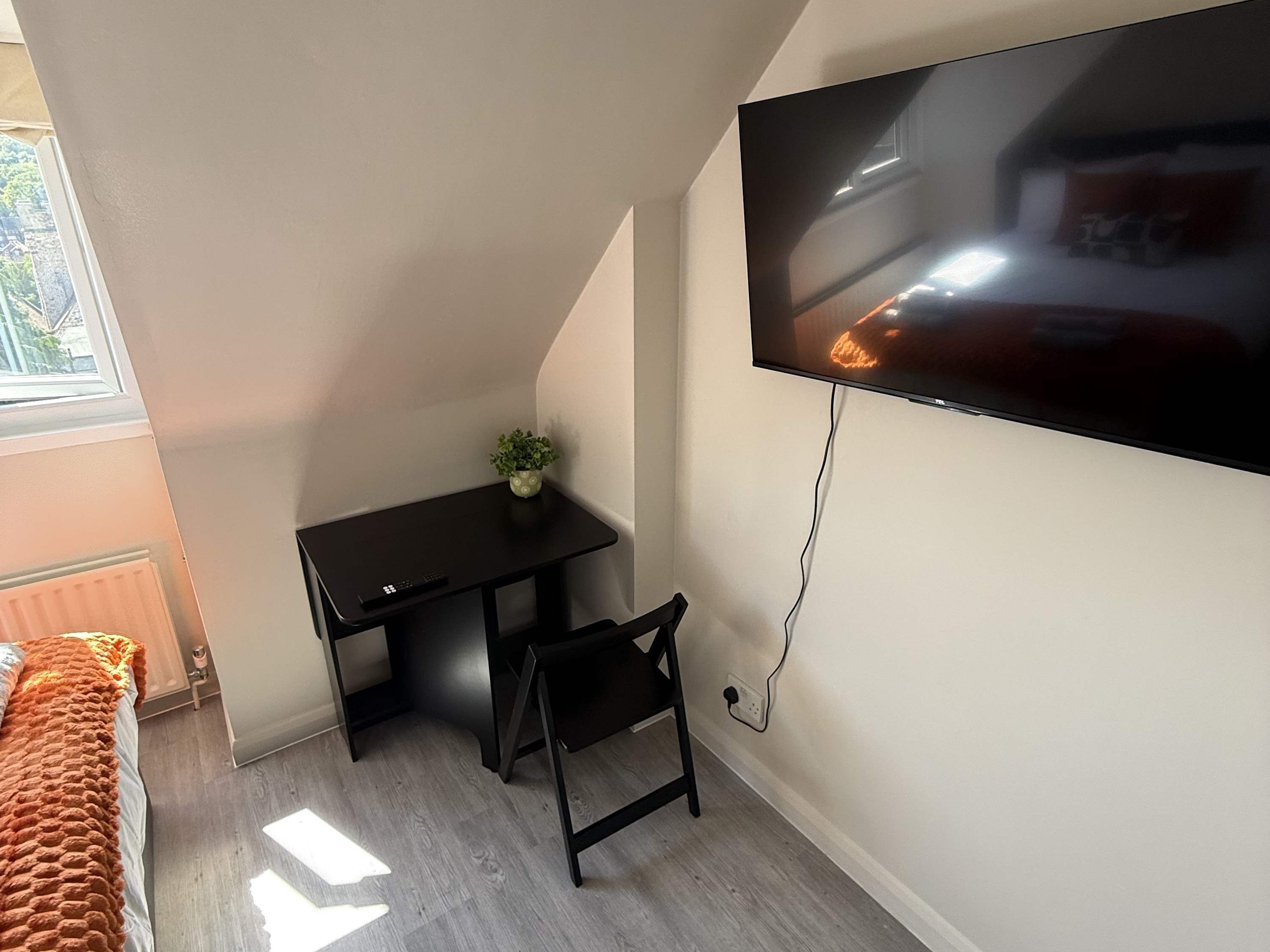 Una habitación con televisión de pantalla plana en la pared, un escritorio pequeño negro con una silla, una planta en maceta y una ventana que proporciona luz natural.