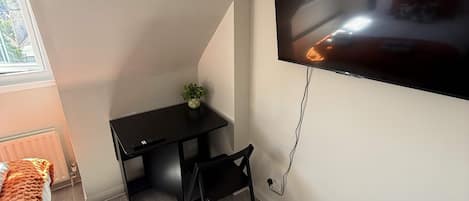 A room with a flat-screen TV mounted on the wall, a small black desk with a chair, a potted plant, and a window providing natural light.