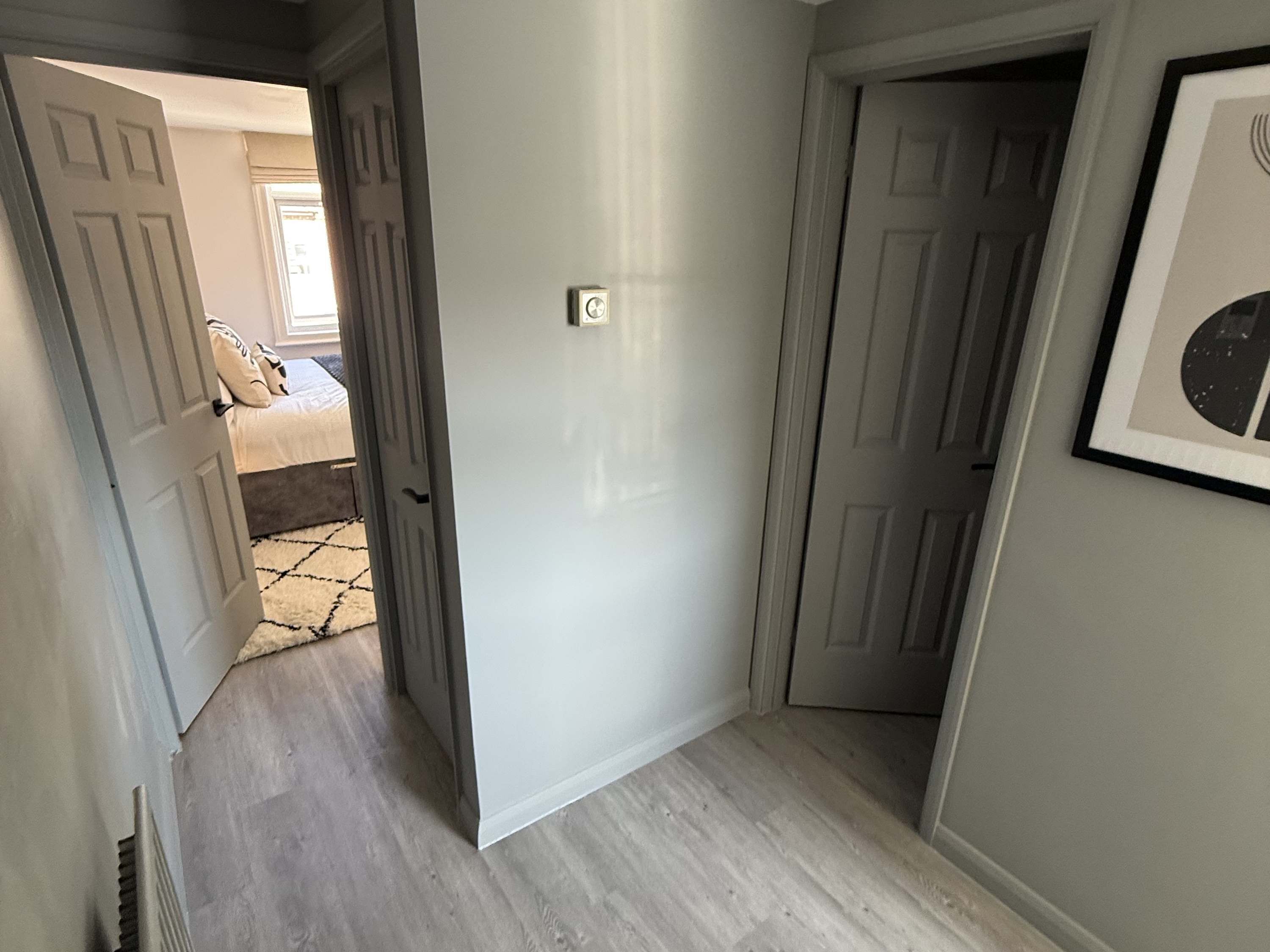 Un couloir avec une porte donnant sur une chambre, un interrupteur et un tableau encadré accroché au mur.