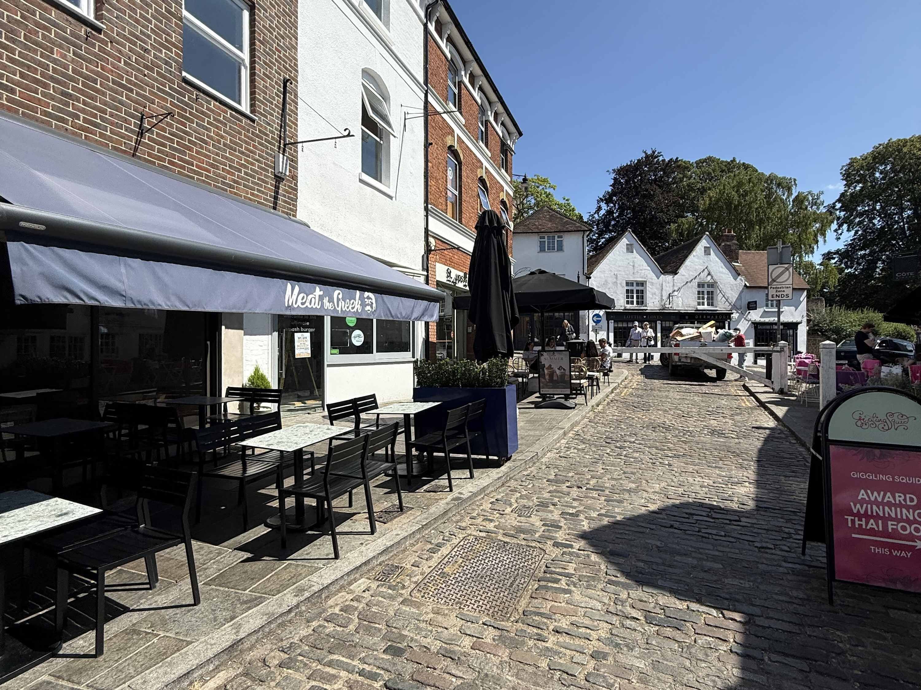 A cobblestone street with outdoor seating at a restaurant named 'Meat the Greek.'