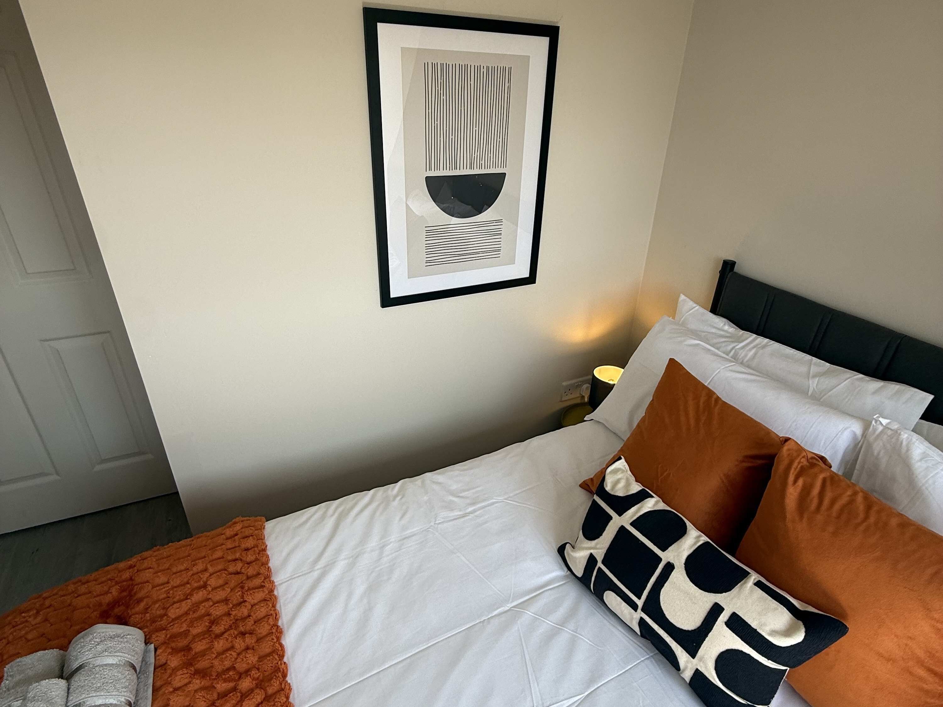 Uma cama bem arrumada com travesseiros brancos e castanhos, uma obra de arte abstrata emoldurada na parede e um abajur.
