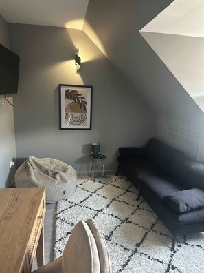 A room with a dark-colored sofa, a small side table with a lamp, a bean bag chair, a framed artwork on the wall, and a wooden table.