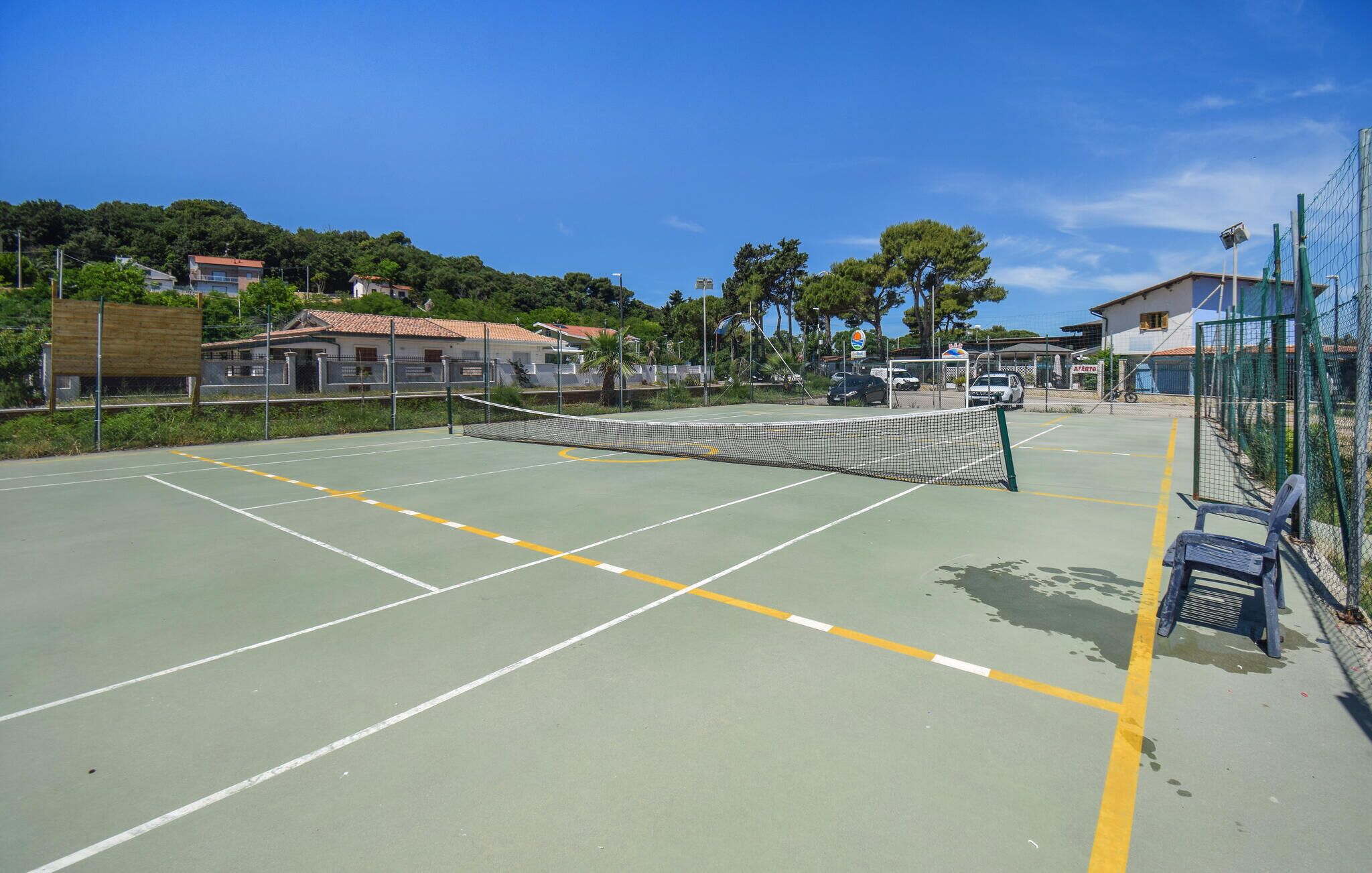 Un campo da tennis all'aperto con una rete, circondato da una recinzione, adiacente a un edificio con un tetto rosso.