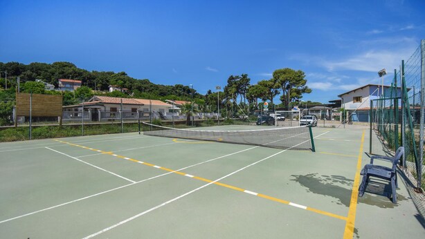 Una cancha de tenis al aire libre con red, rodeada por una cerca, contigua a un edificio de techo rojo.
