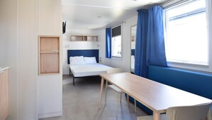 Habitación de hotel pequeña con una cama individual, una mesita y una silla. Cuenta con una ventana con cortinas, un estante y una lámpara fijada a la pared.