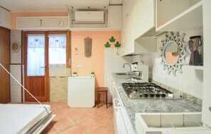 Uma cozinha compacta com fogão a gás, pia e geladeira. Há um banquinho de madeira e um vaso de planta no balcão.