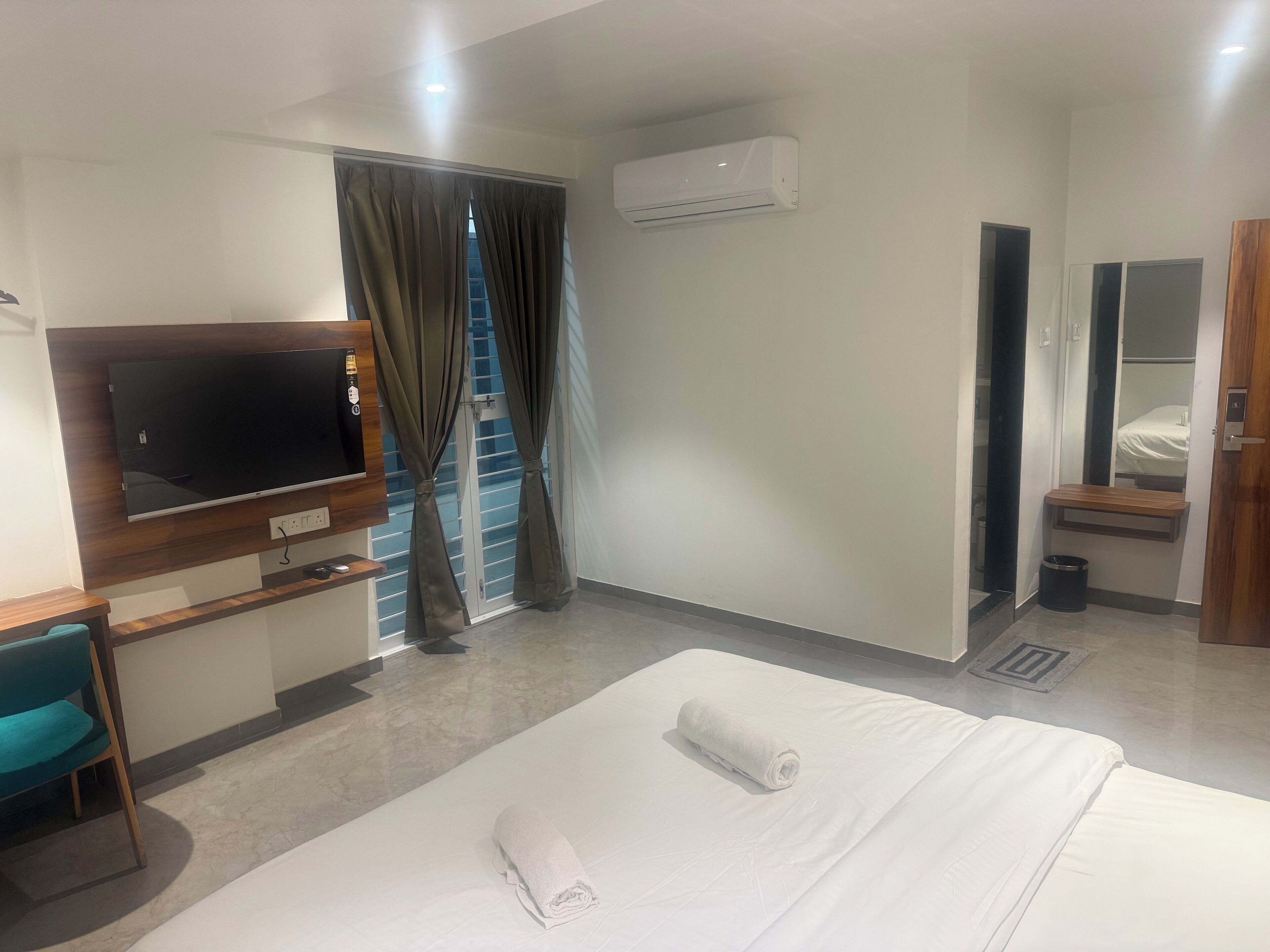 Habitación de hotel con cama, televisión, ventana con cortinas, aire acondicionado y un pequeño estante de madera.