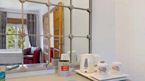 Una habitación de hotel con ventana, una mesa con hervidor, tazas y un pequeño cuadro.