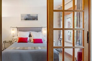 Un dormitorio con cama, almohadas, mesita de noche, lámpara de pared y ventana con vistas al exterior.