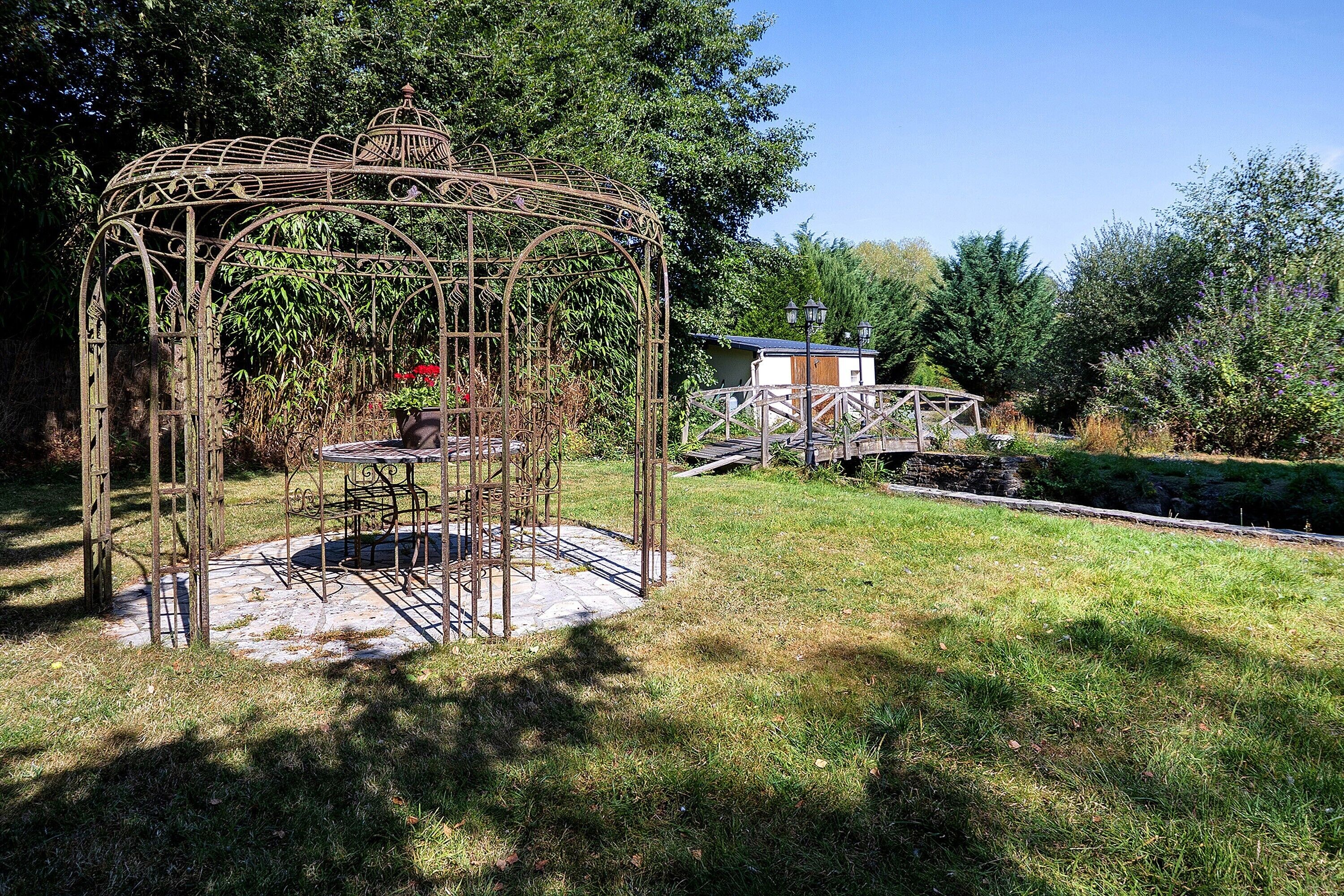 A gazebo with a circular base and intricate metalwork, surrounded by a grassy area and trees.