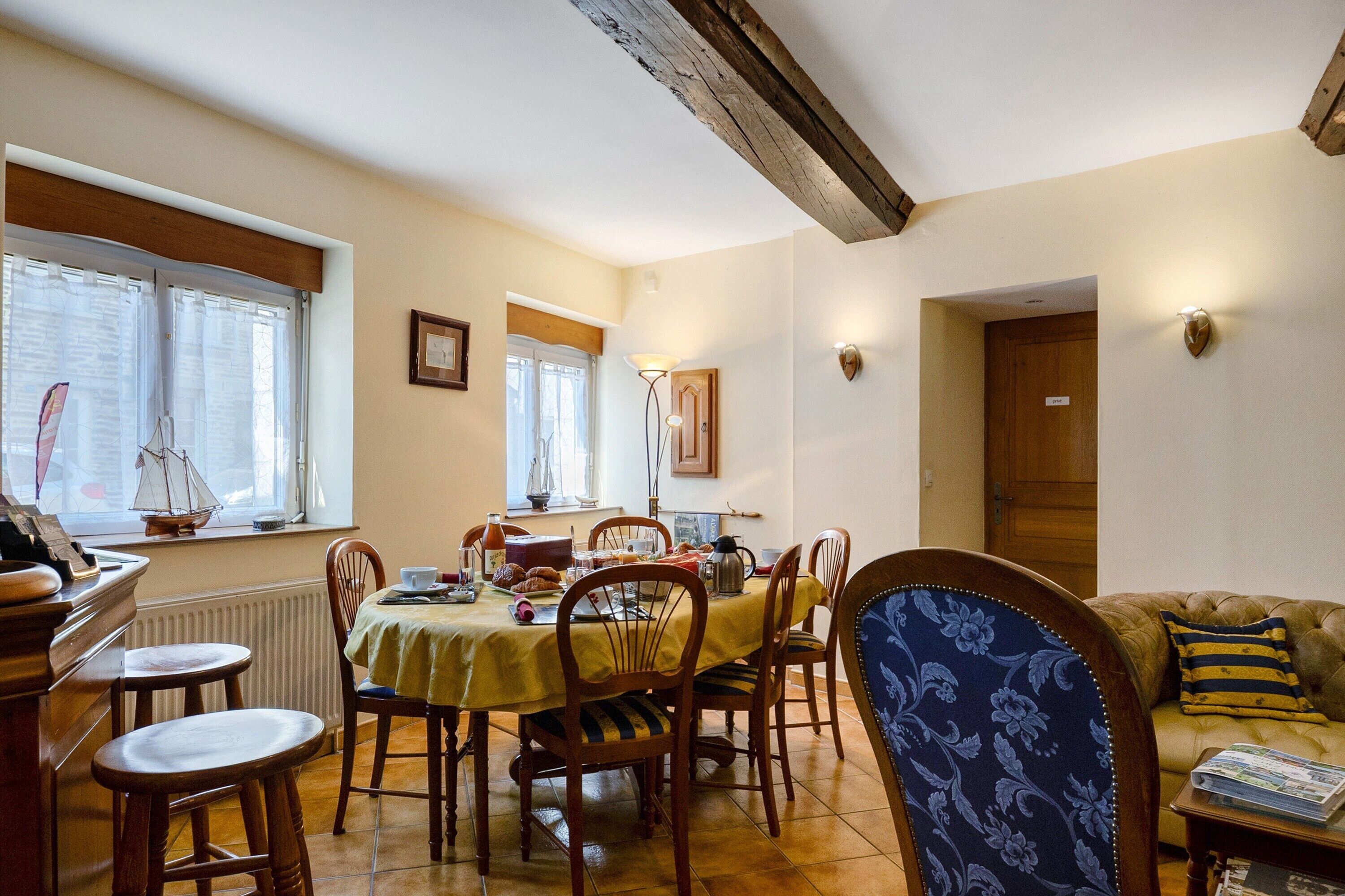 Un coin repas avec une table dressée pour quatre personnes, des chaises, une fenêtre avec des rideaux, un radiateur et une poutre en bois au plafond.