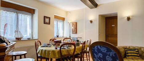 Un coin repas avec une table dressée pour quatre personnes, des chaises, une fenêtre avec des rideaux, un radiateur et une poutre en bois au plafond.