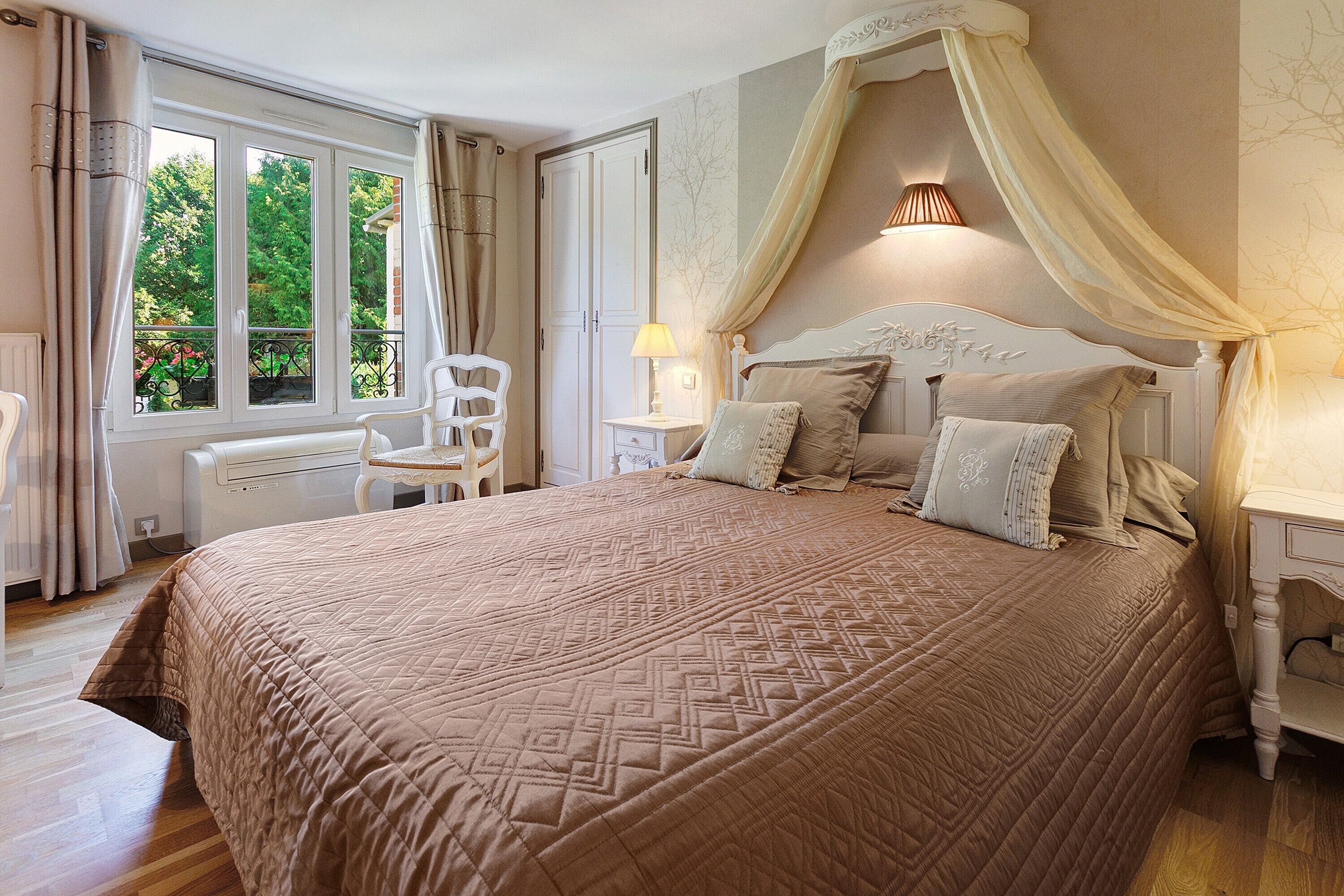 A bedroom with a large bed, a chair, a bedside table, a lamp, and a window with curtains.