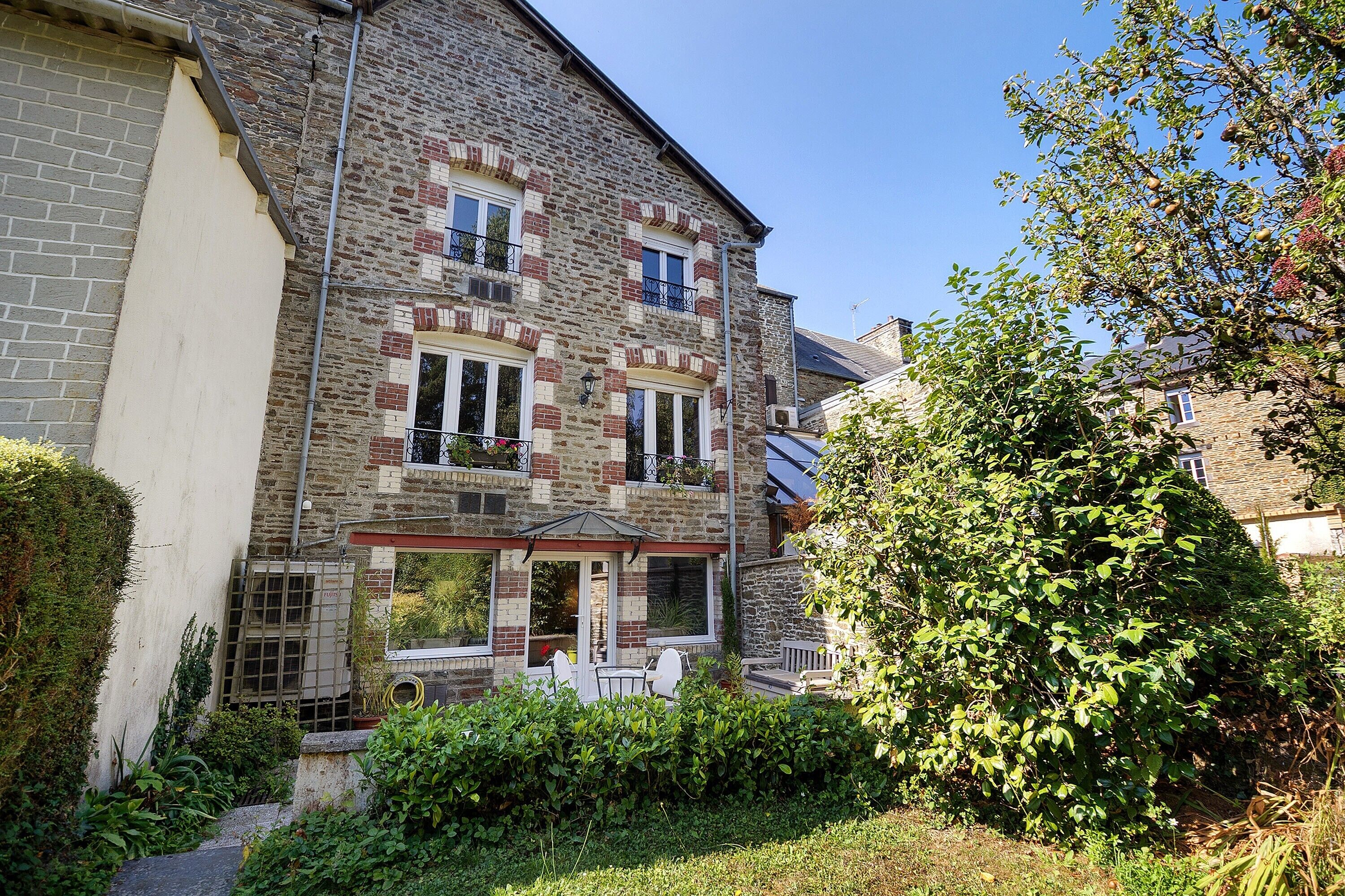 A two-story stone building with a balcony, a small garden, and a tree.