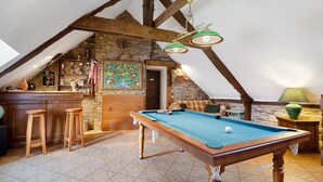 A cozy attic room with a pool table, wooden stools, a bar area, and a stone wall.