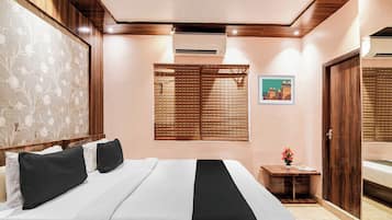Una habitación de hotel con cama, ventana con persianas, una mesita con un florero y una puerta.