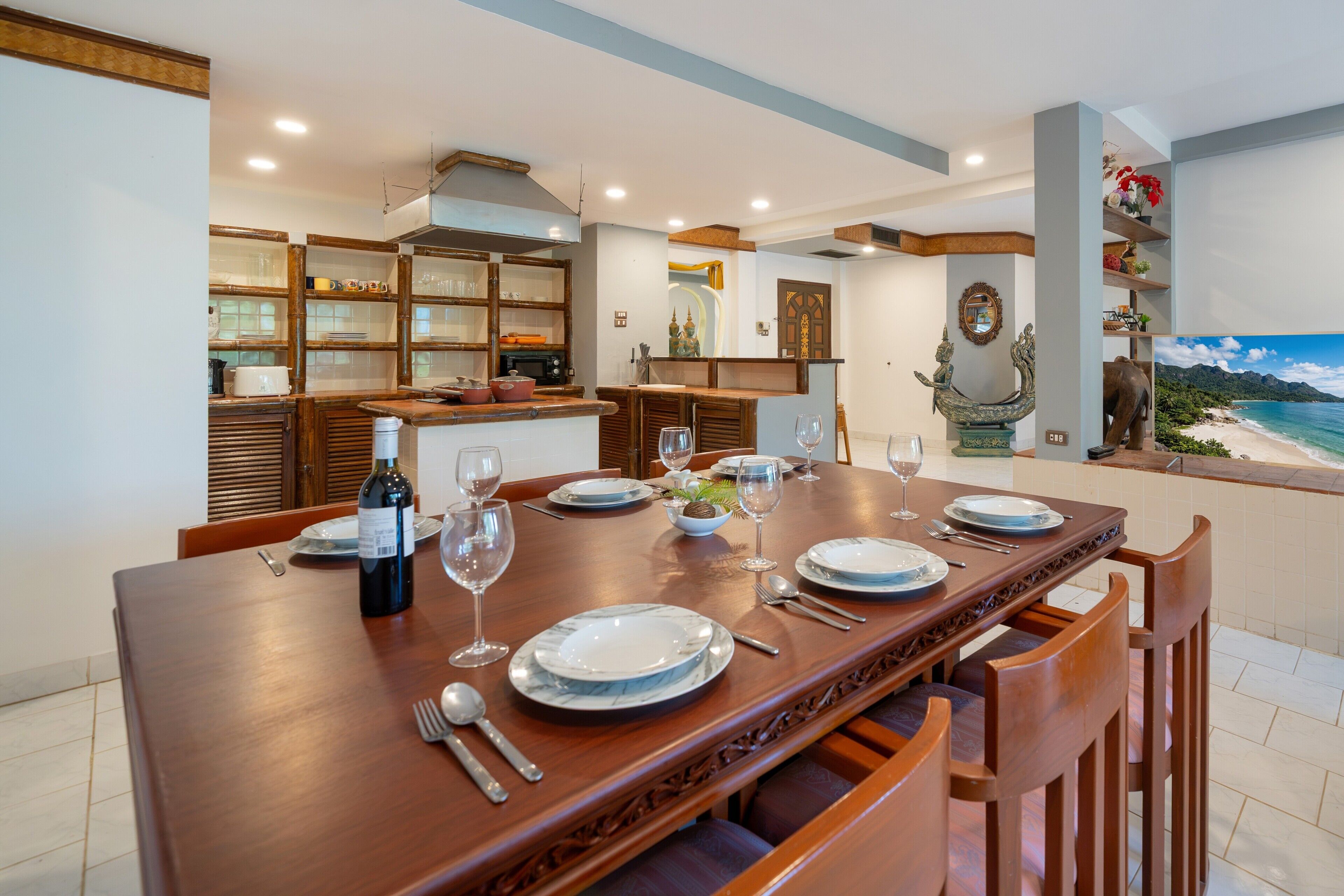 Comedor con una larga mesa de madera preparada para una comida, una cocina al fondo y vista a una playa por una ventana.