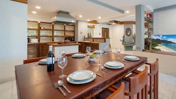 A dining area with a long wooden table set for a meal, a kitchen in the background, and a view of a beach through a window.