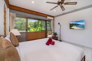 Habitación de hotel con cama, ventilador de techo, ventana con cortinas, mesita de noche, lámpara y un cuadro de playa.